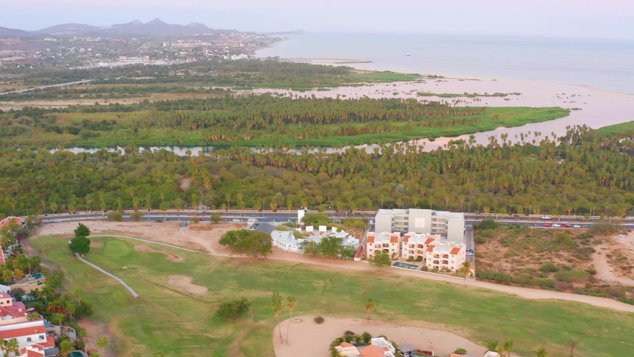 Opening aerial frame in San Jose del Cabo showing golf greens and paths in front, a road and residential buildings behind, then trees, sand, ocean, hazy hills, and no visible people.