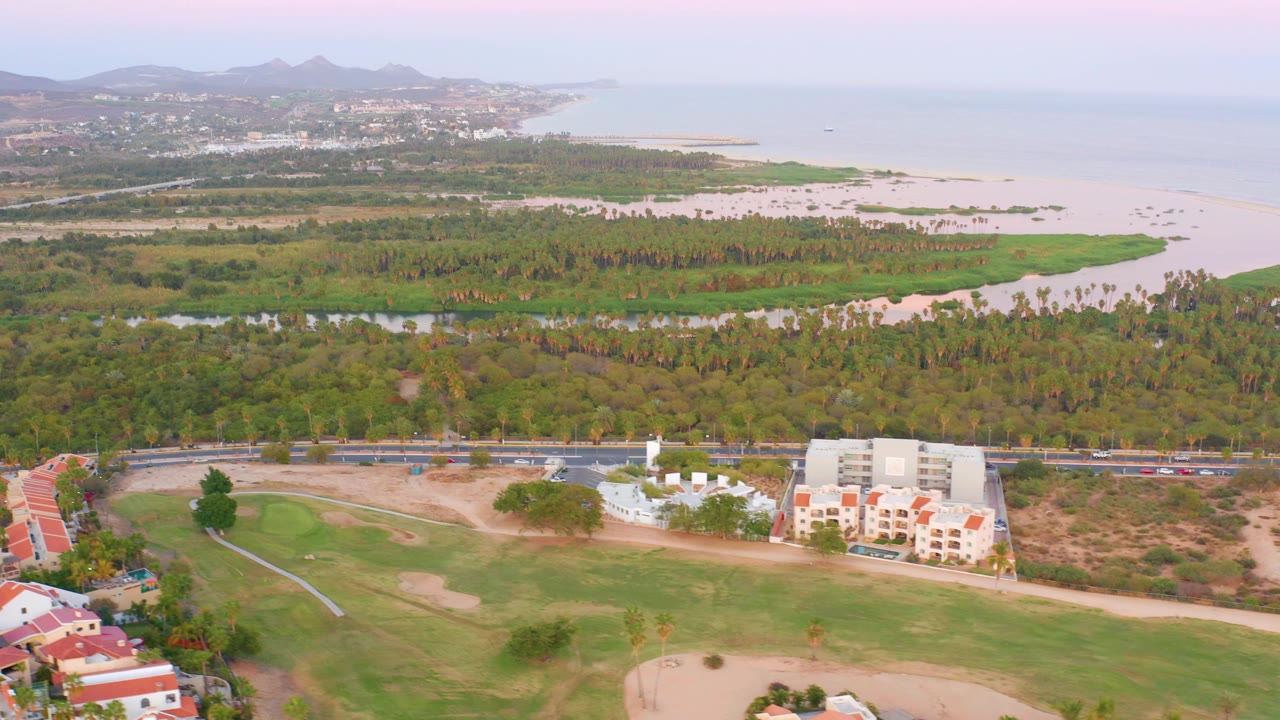 San Jose del Cabo aerial frame with a large fairway at lower left, white residential buildings near the road, a green belt, pale sandy coastline, ocean horizon, and no visible people.