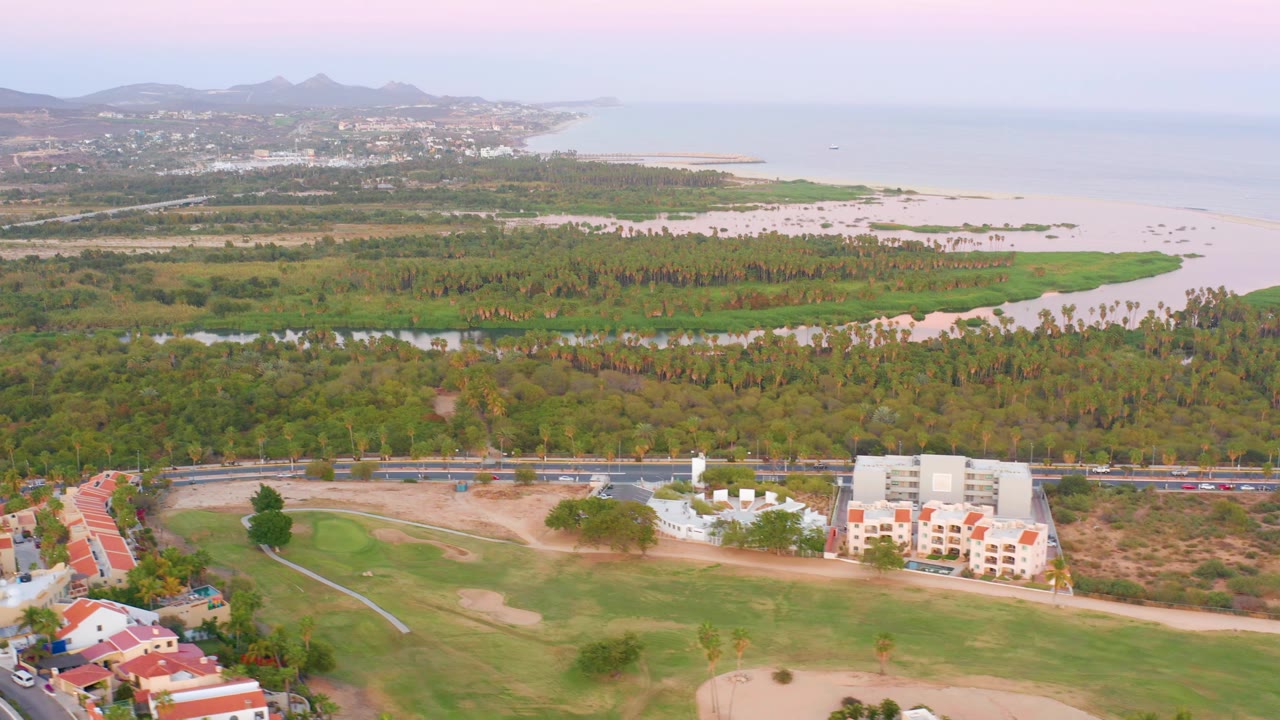 Mid-clip San Jose del Cabo view showing the golf course shifted across the lower frame, clustered buildings by the road, dense greenery, exposed sand, ocean water, distant hills, and no visible people.
