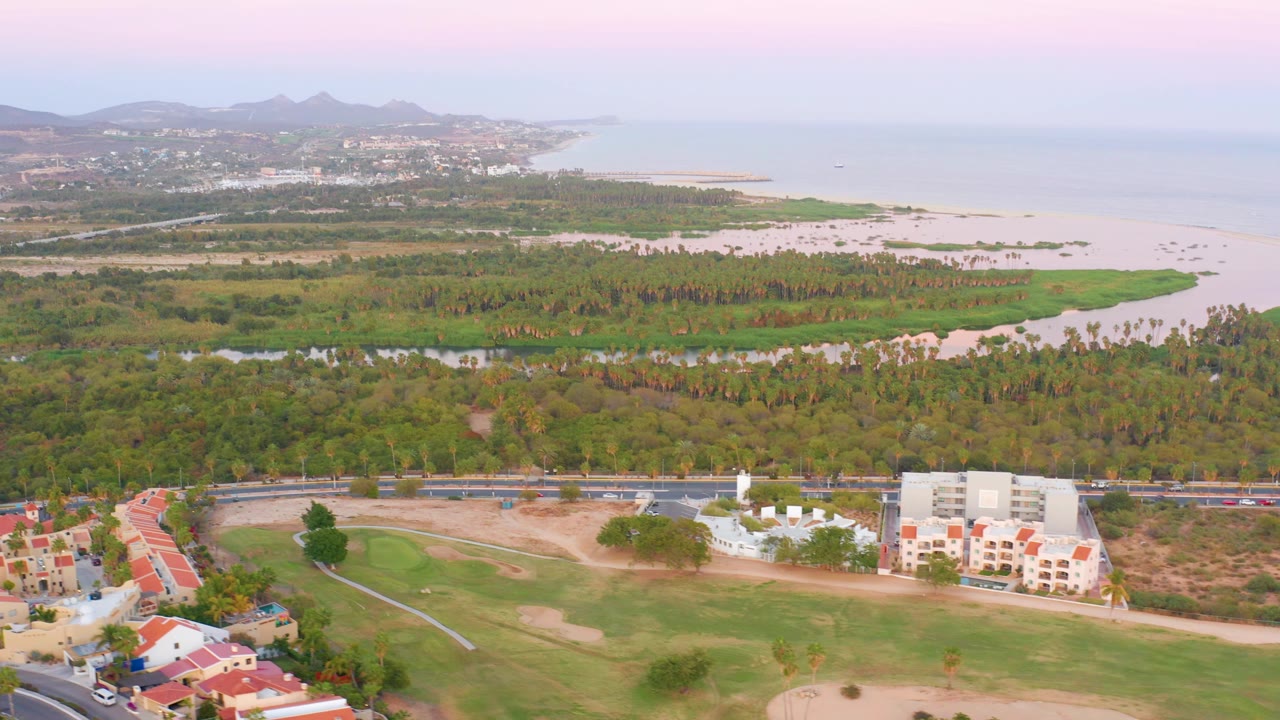 Aerial view of resort-style homes, golf course, road, dense trees, sandy coast, ocean, and hills.