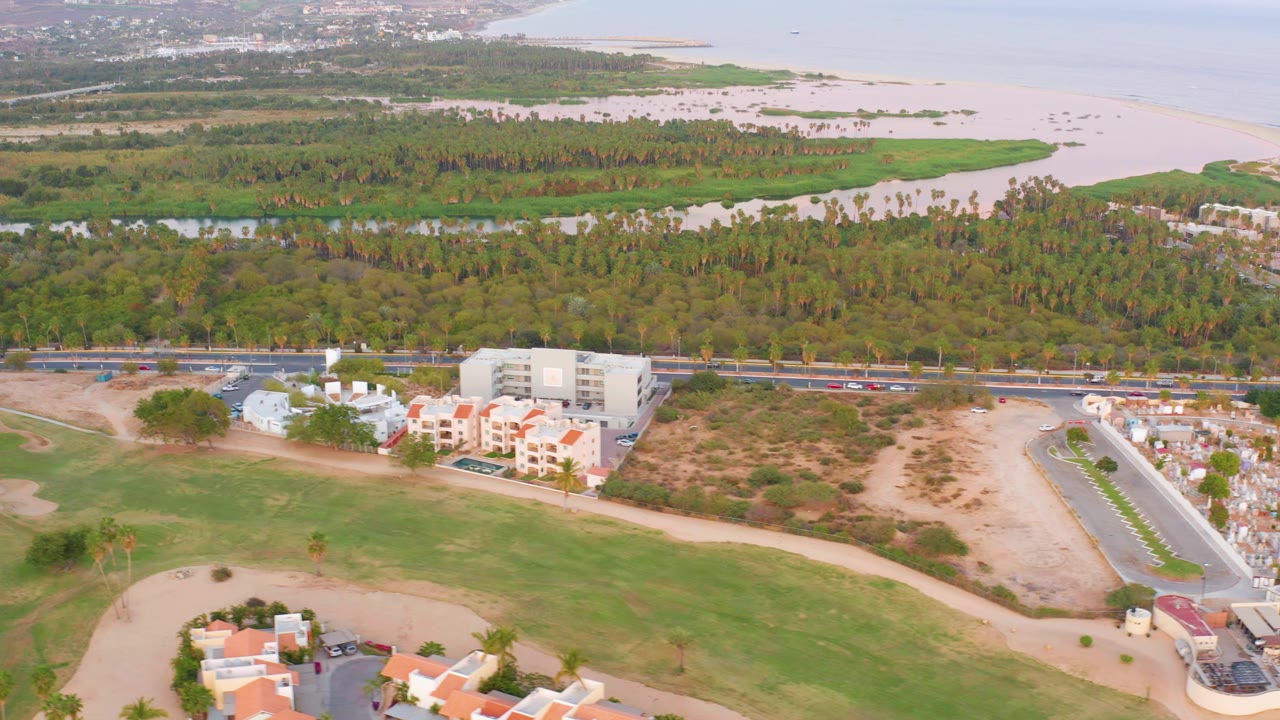 Aerial frame over San Jose del Cabo showing golf course fairways and sand traps in front, residential buildings and a road beyond, tree cover and pale ocean in the distance, with no visible people.