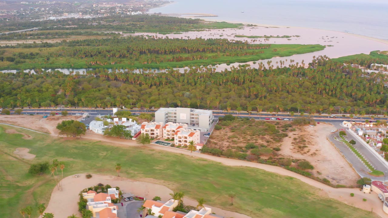 Second aerial frame in San Jose del Cabo with broad green fairways across the lower half, white and terracotta-roofed residences near a road, dense vegetation, and distant sea, with no visible people.