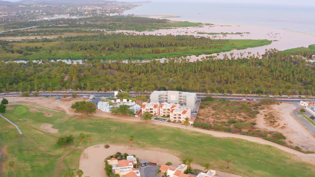 Wide drone view of fairway, sand trap, buildings, road, tree canopy, and shoreline.