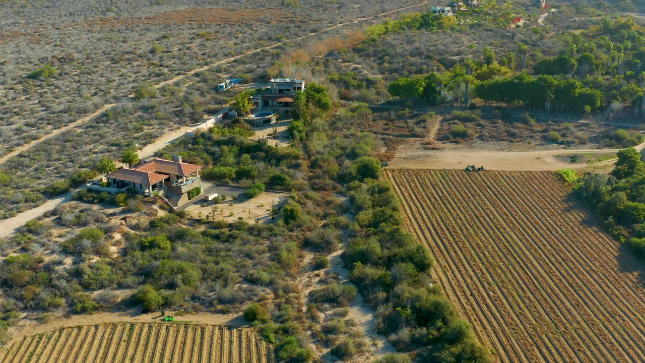 Aerial frame over Todos Santos, Baja California Sur showing a red-roof rural house near plowed fields, dry scrub, a dirt road, scattered greenery, and no visible people.