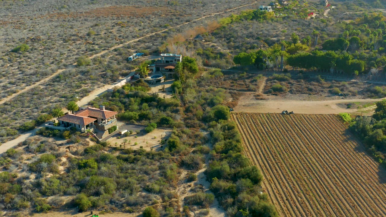 Aerial view in Todos Santos, Baja California Sur with the rural house lower left, a dirt road crossing the dry land, plowed field rows to the right, vehicles, greenery, and no visible people.