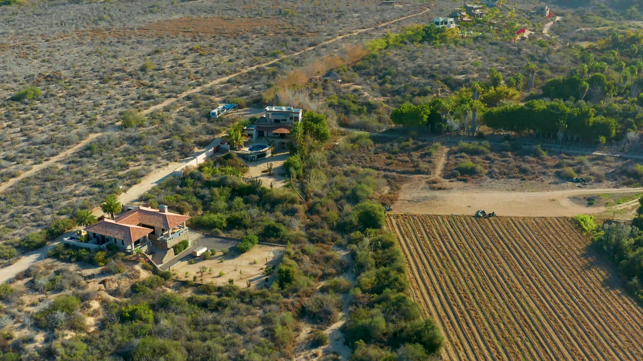 Aerial frame over Todos Santos, Baja California Sur showing the house and access road left of center, long brown field rows to the right, scrub-covered slopes, vehicles, and no visible people.