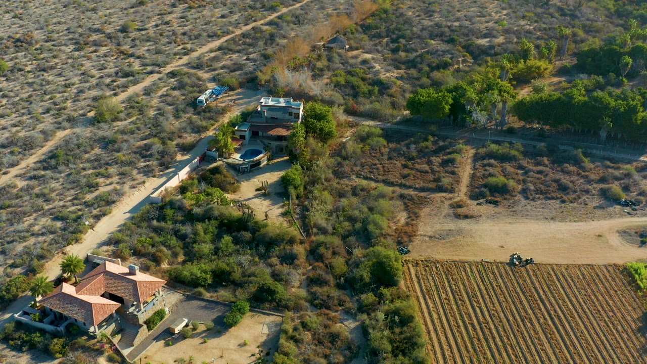 Aerial frame over rural Todos Santos, Baja California Sur, showing red-roofed houses, scrubby dry land, a dirt access path, a blue vehicle, a plowed field, and a tiny person near the path.