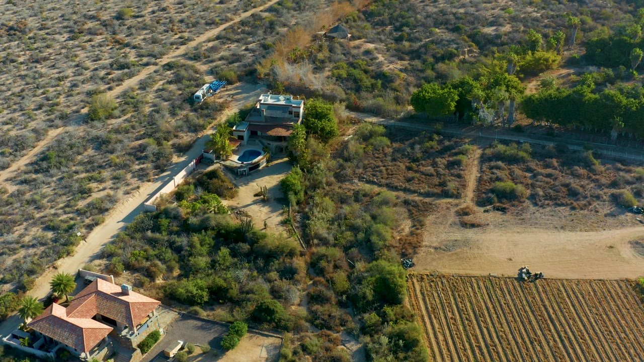 Drone view of a rural house compound beside dry brush and field rows.