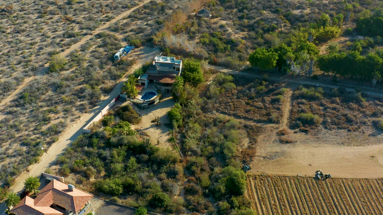 Aerial frame over Todos Santos, Baja California Sur, showing the house cluster left of center, a plowed field along the lower right, scrub-covered dry land above, a blue vehicle, and no clearly visible people.
