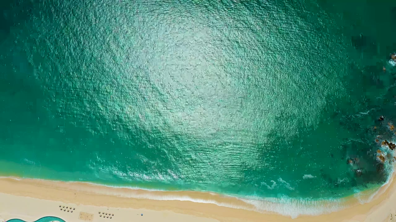 Top-down view at Marquis Los Cabos, Mexico, showing bright turquoise water, pale sand along the bottom edge, dark submerged rocks at right, and no visible people.