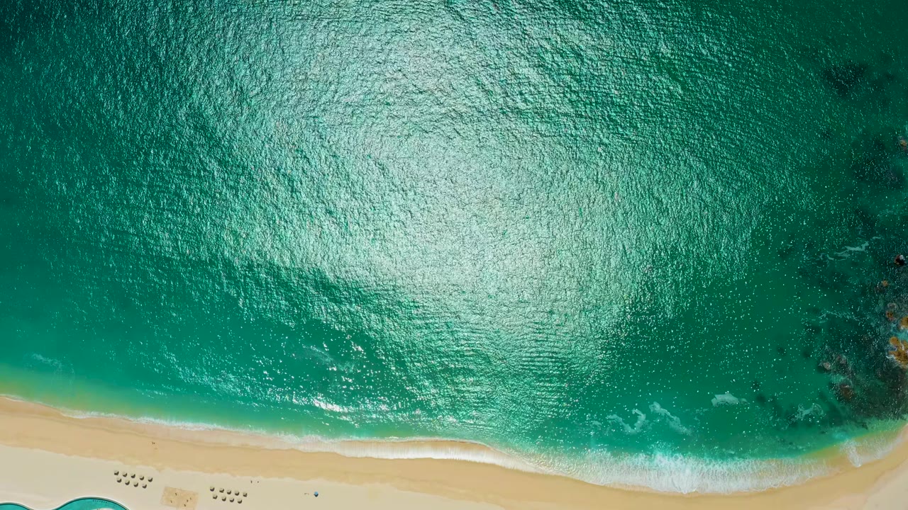 Top-down view at Marquis Los Cabos, Mexico, with turquoise water, sun glare, a pale sandy shoreline, small foam lines, and no visible people.