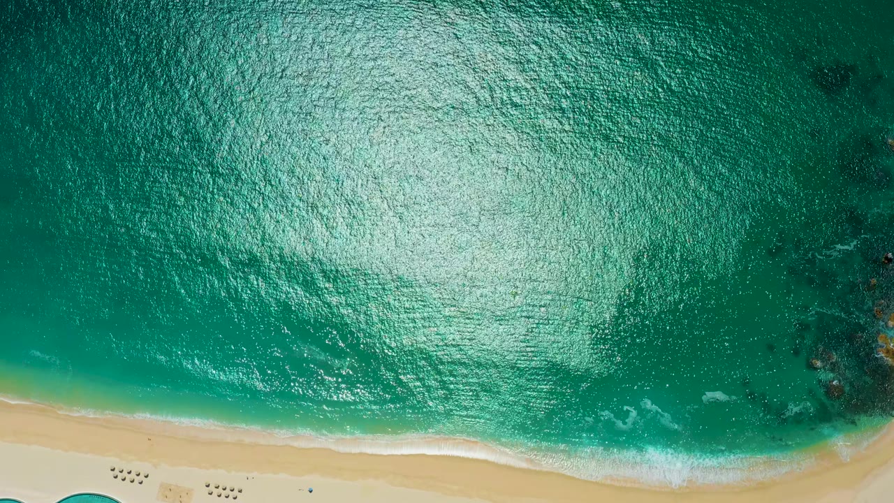 Overhead frame from Marquis Los Cabos, Mexico, showing bright reflected light on turquoise water, a narrow surf line, pale sand, and no visible people.