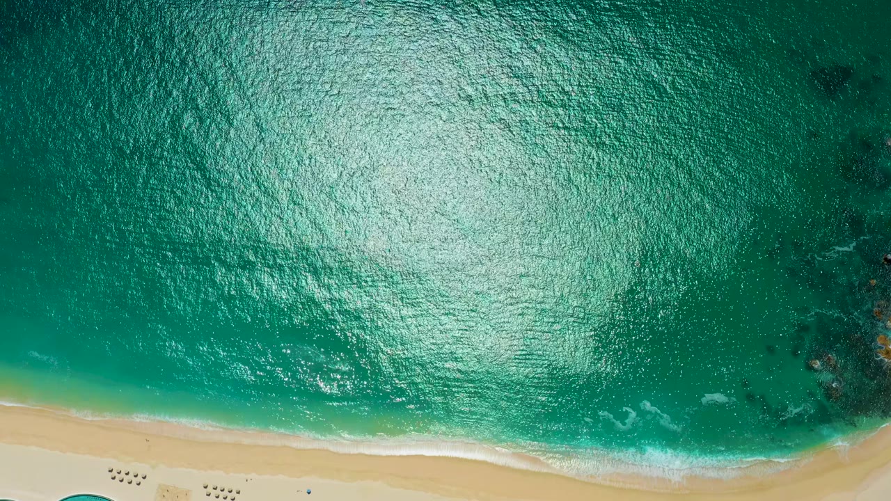 Aerial top-down view at Marquis Los Cabos, Mexico, with a broad patch of sun glare across green-blue water, soft foam near the sand, and no visible people.
