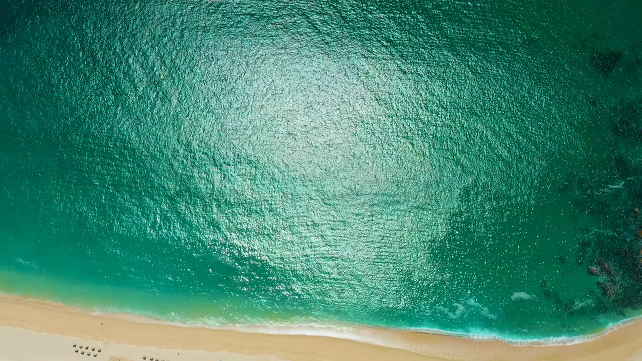 Overhead view of turquoise ocean water meeting sand at Marquis Los Cabos, with bright sun reflection on the rippled surface and no visible people.