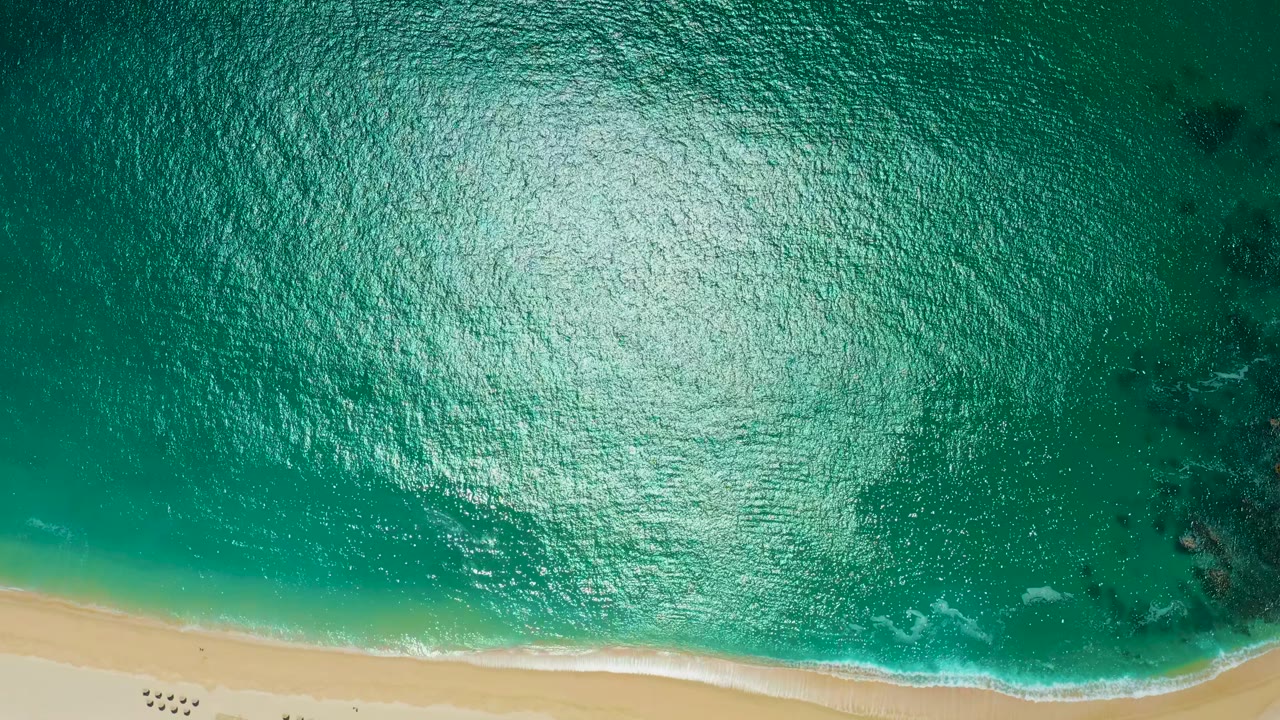 Aerial frame at Marquis Los Cabos showing teal water, a narrow strip of sand along the bottom edge, small white foam lines, and no visible people.