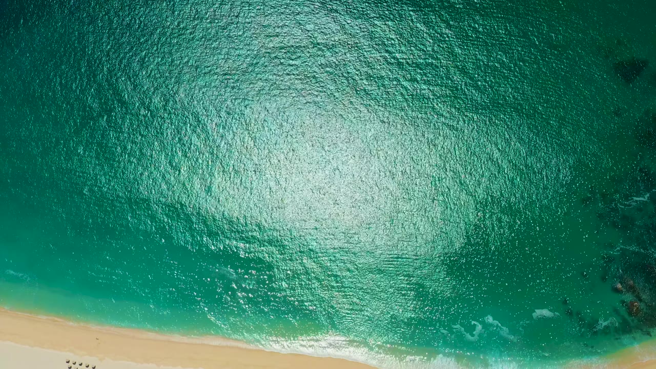 Overhead view at Marquis Los Cabos, Mexico, showing turquoise rippled water, a strip of sand at the bottom, small people walking on the beach, and white foam touching the shore.