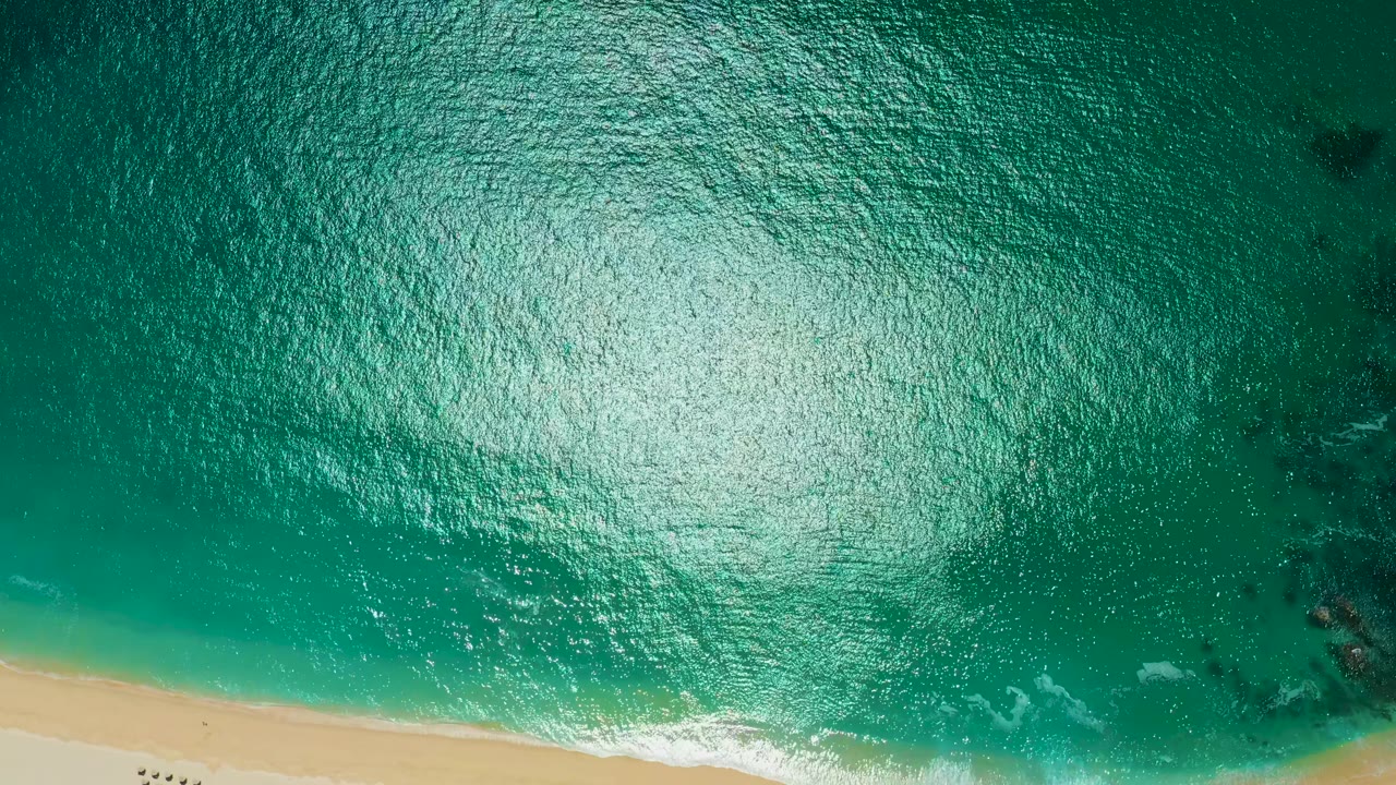 Bright turquoise water with sun glare, beach along the bottom, and dark patches near shore.