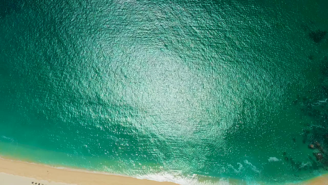 Top-down Marquis Los Cabos, Mexico beach view with turquoise water across most of the frame, a sandy strip at the bottom, broken white foam at the waterline, and small figures on the sand.