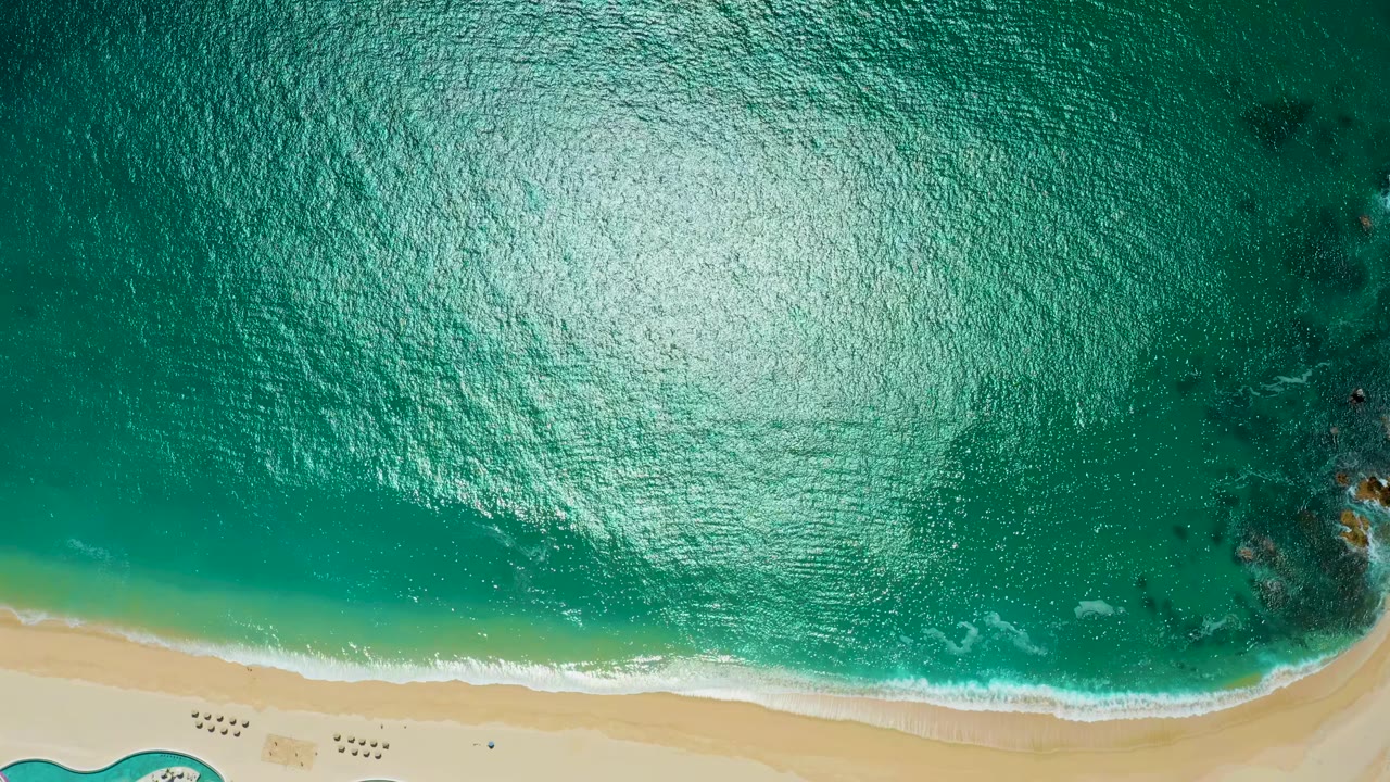 Aerial view at Marquis Los Cabos, Mexico, showing turquoise water, bright surface reflection, a pale sandy shoreline, small foam lines, and no visible people.
