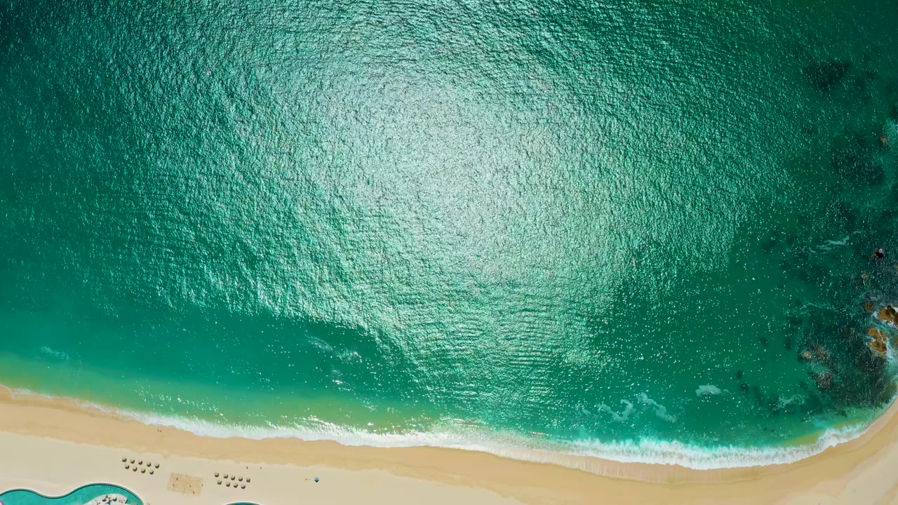 Aerial frame over Marquis Los Cabos, Mexico, with rippled turquoise ocean, a bright reflected patch on the water, foamy surf touching sand, and no visible people.
