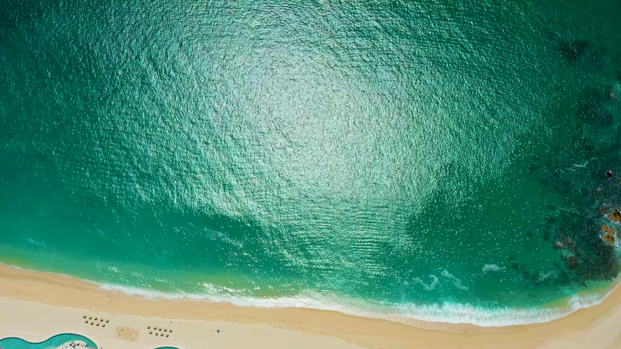 Top-down view at Marquis Los Cabos, Mexico, where dark offshore rock patches sit in turquoise water above a narrow foam band and pale sand, with no visible people.