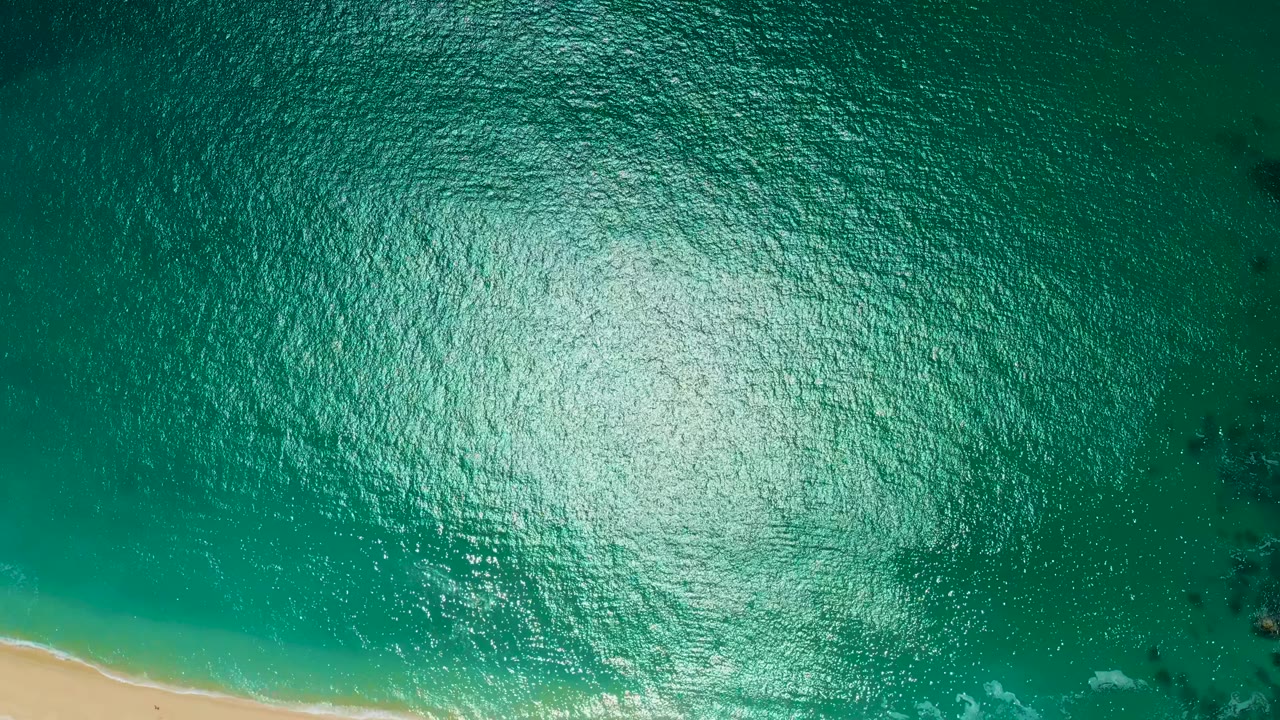 Aerial frame at Marquis Los Cabos, Mexico, with green-blue rippled water, a large reflected light patch, sand entering from the lower left, and no visible people.