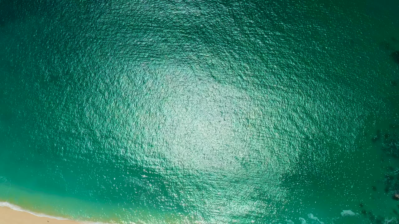 Overhead Marquis Los Cabos, Mexico frame showing turquoise water with a pale sand strip at the lower left, scattered foam near shore, dark rocks to the right, and no visible people.