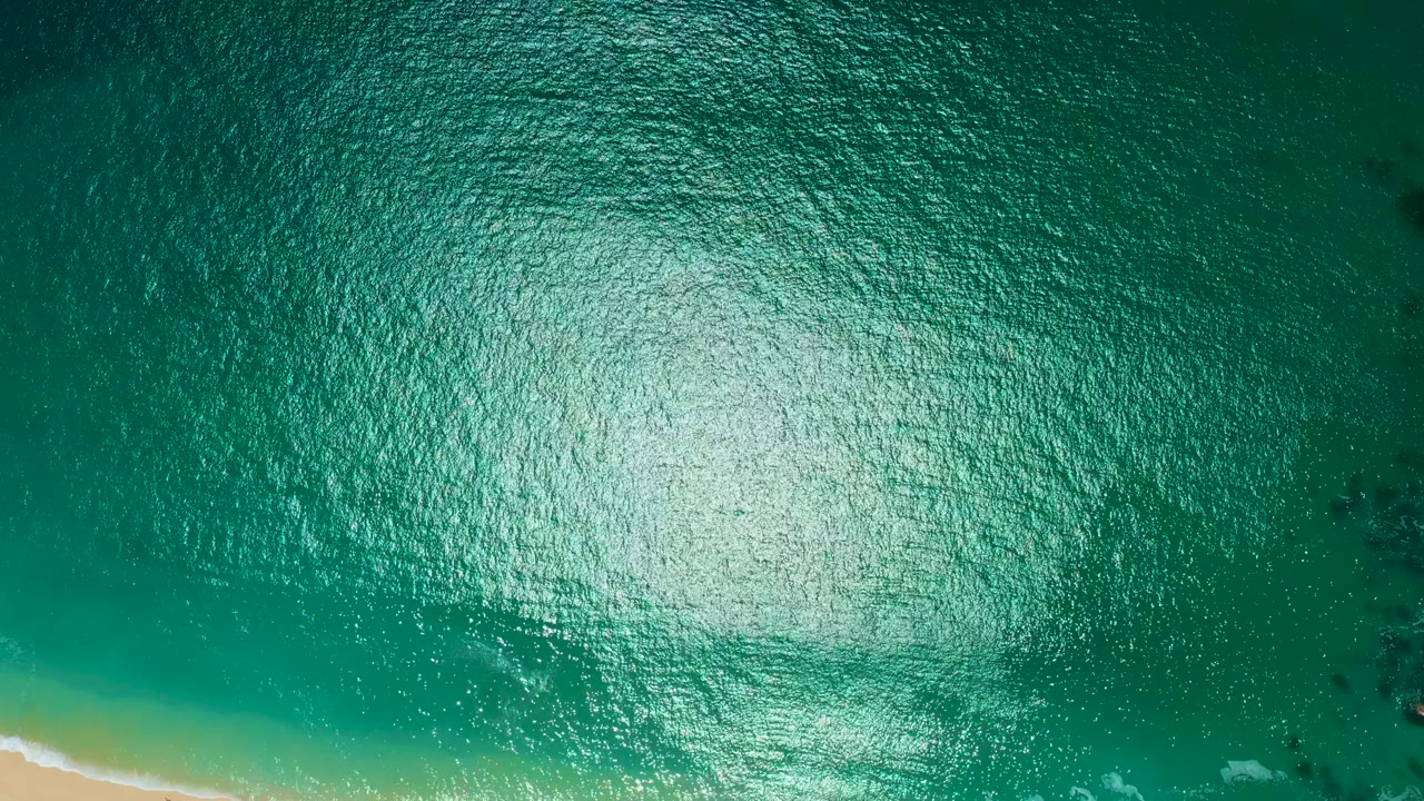 Aerial view at Marquis Los Cabos, Mexico, with bright glare across rippled green water, a sandy shoreline along the lower left corner, faint foam, and no visible people.