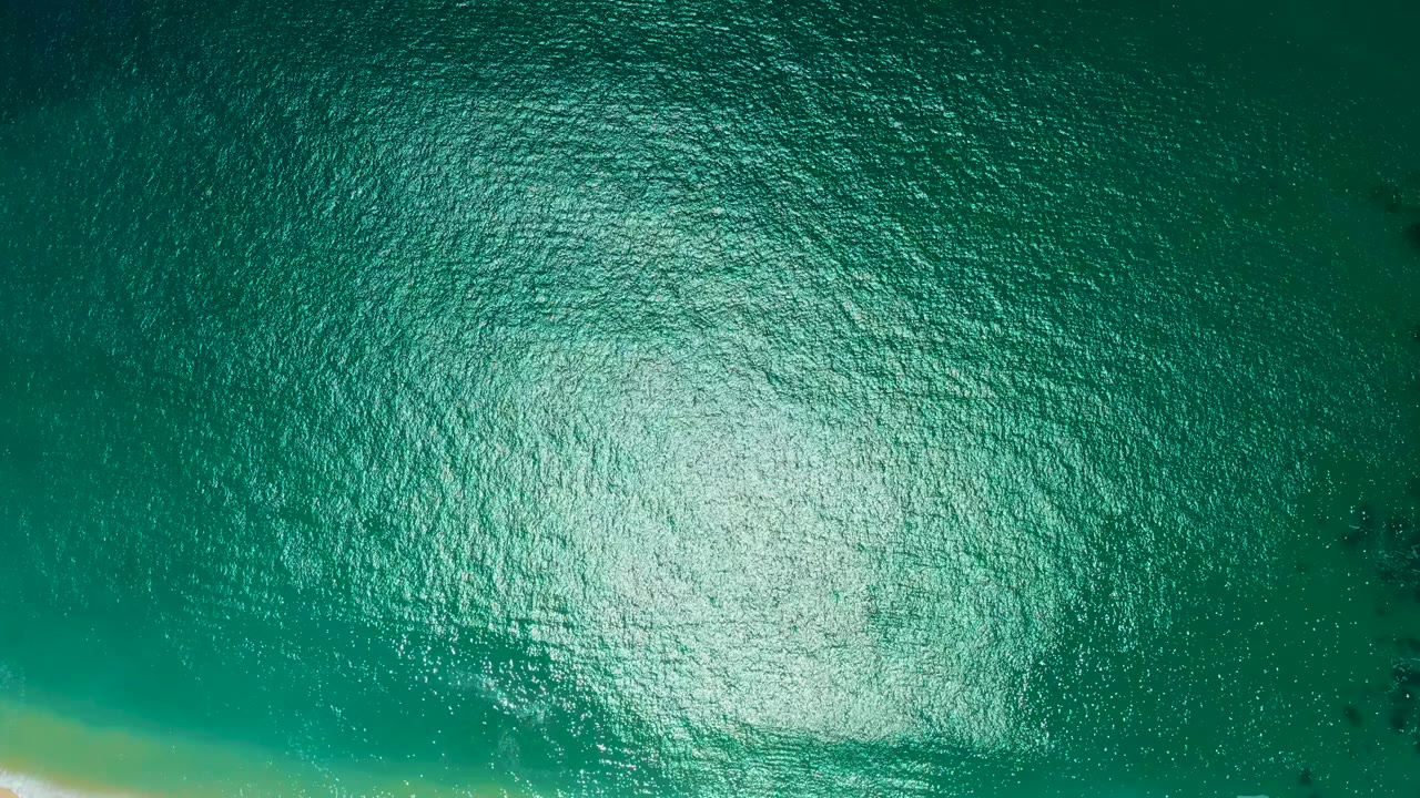 Overhead view of teal ocean water at Marquis Los Cabos, Mexico, with a broad bright reflection near the lower left and no visible people.