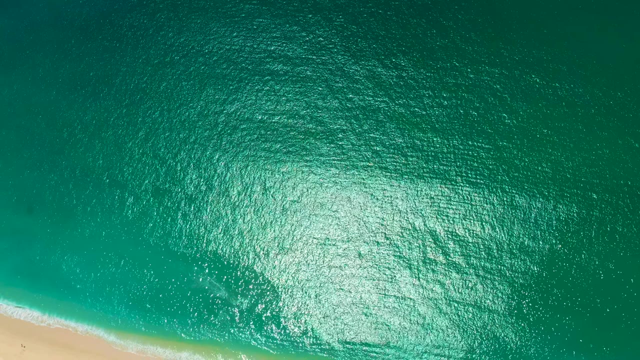 High aerial view at Marquis Los Cabos, Mexico, with turquoise rippled water, bright sun reflection, a thin surf line, and a narrow strip of pale sand at the bottom edge.