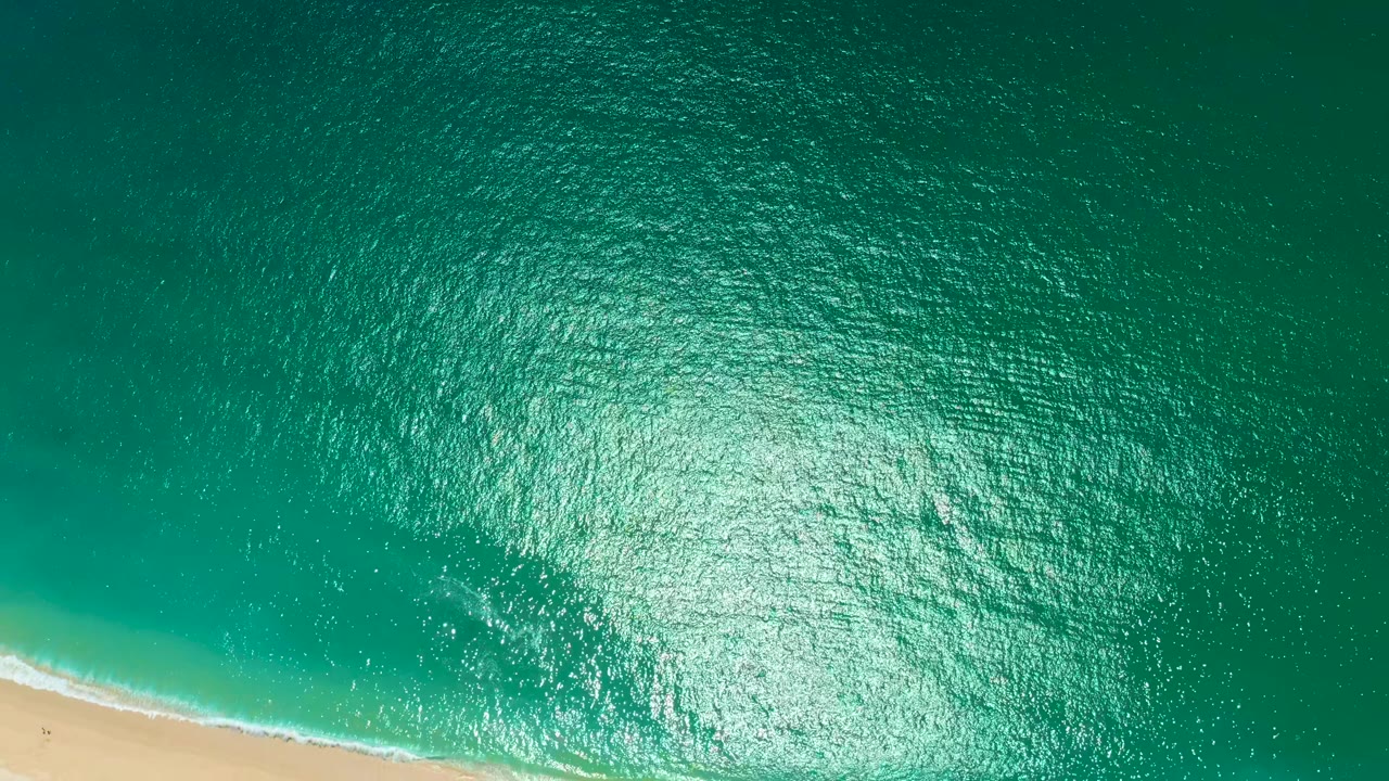 Aerial view at Marquis Los Cabos, Mexico, with the sunlit ocean dominating the frame, a diagonal strip of sand along the lower left, and a narrow white break of wave foam near shore.