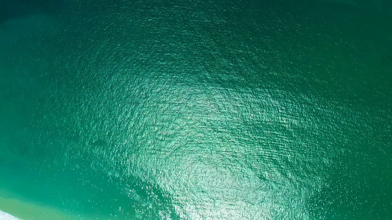 Overhead aerial view of turquoise-green ocean water with a narrow strip of sandy beach at the bottom-left edge