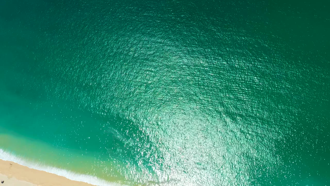 Overhead view at Marquis Los Cabos, Mexico, with turquoise water, bright surface shimmer, a narrow sandy shoreline at lower left, and no visible people.