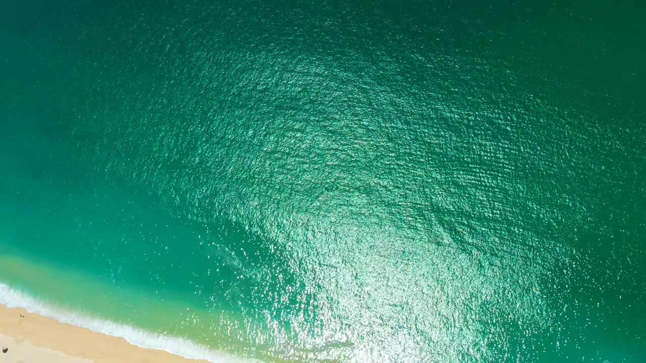 Overhead Marquis Los Cabos frame with green-blue water, a stronger band of white foam along the sand, bright water glare near center, and no visible people.