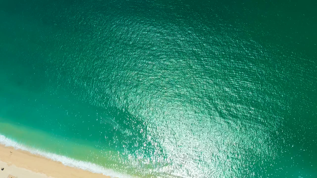 Overhead view from Marquis Los Cabos with turquoise surf angled across the lower left shoreline, dense ripples in the upper water, and no visible people.