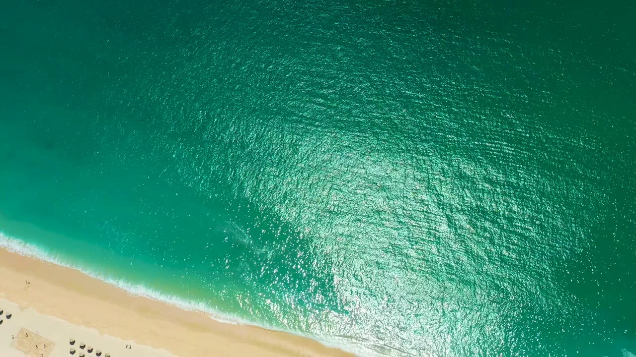Aerial view at Marquis Los Cabos, Mexico, with turquoise water filling most of the frame, pale sand at the lower edge, small beach umbrellas, and no visible people.