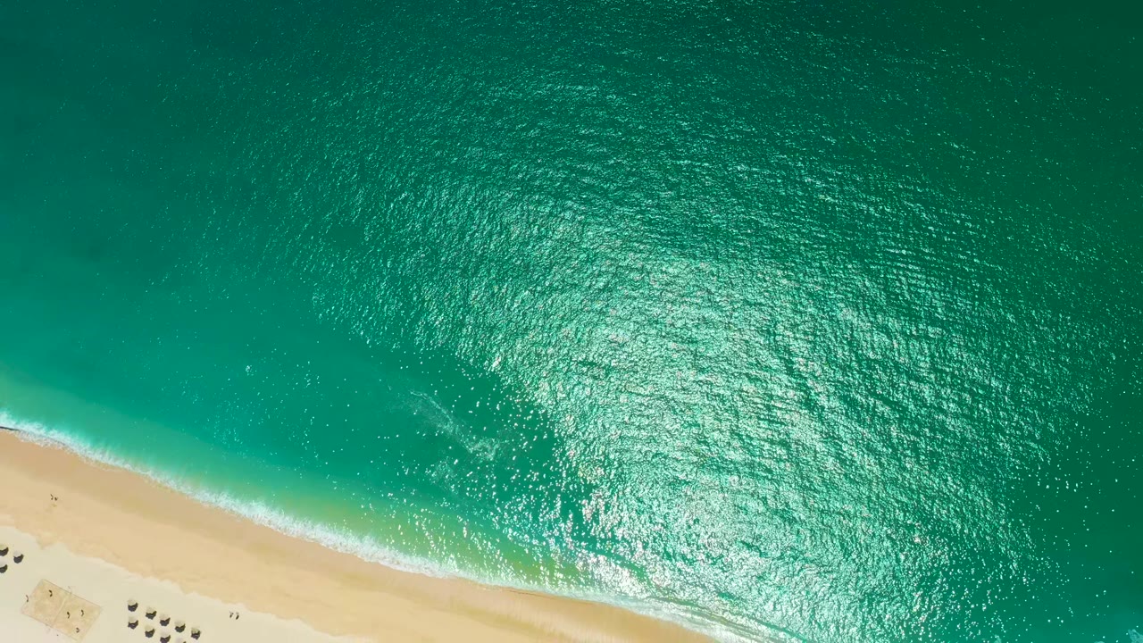 Aerial frame from Marquis Los Cabos, Mexico, showing bright sun glitter across rippled turquoise water and a narrow white foam line along the sand, with no visible people.
