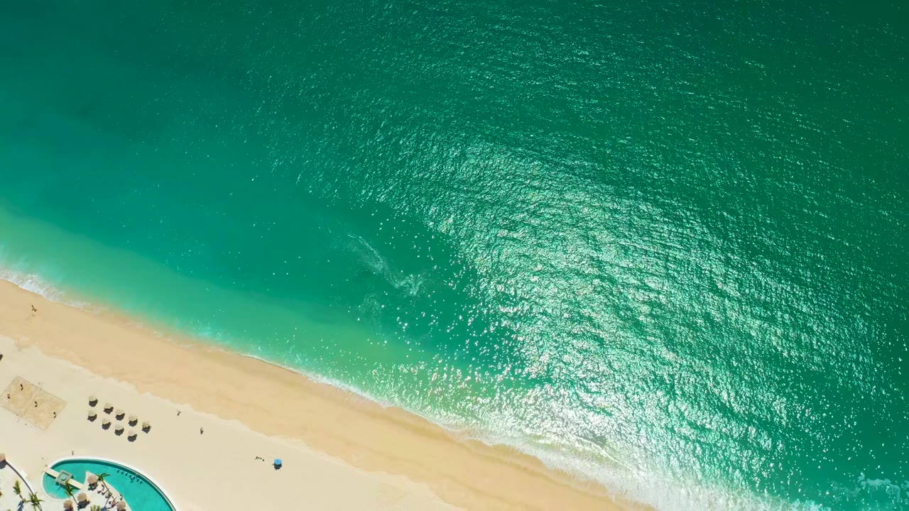 Turquoise water and pale sand shoreline with umbrellas and a pool corner below.
