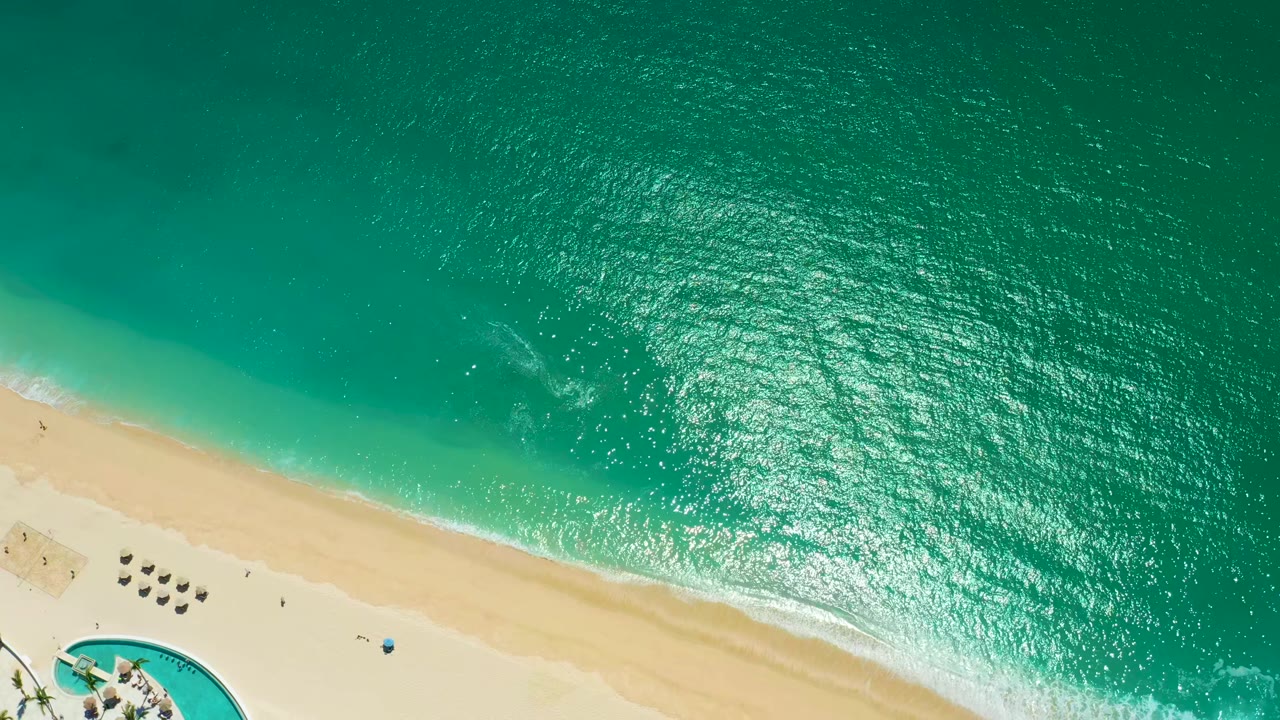 Overhead frame at Marquis Los Cabos, Mexico showing a wide field of turquoise ocean, a narrow foam line at the sand, clustered beach umbrellas, pool edge, and tiny visible people on the beach.