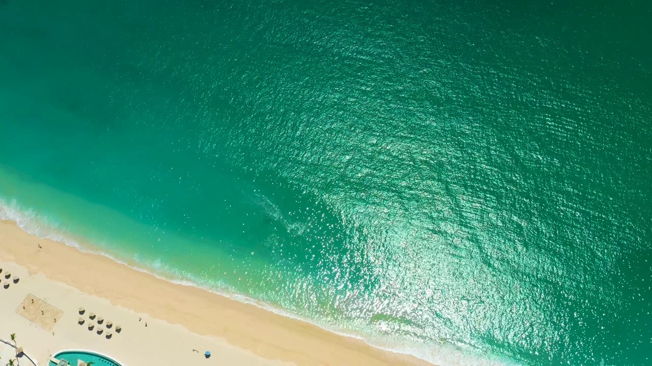 Aerial frame from Marquis Los Cabos, Mexico, showing the foam line shifted along the pale sand, beach umbrellas grouped near the lower left, bright sun reflection on the water, and no visible people.