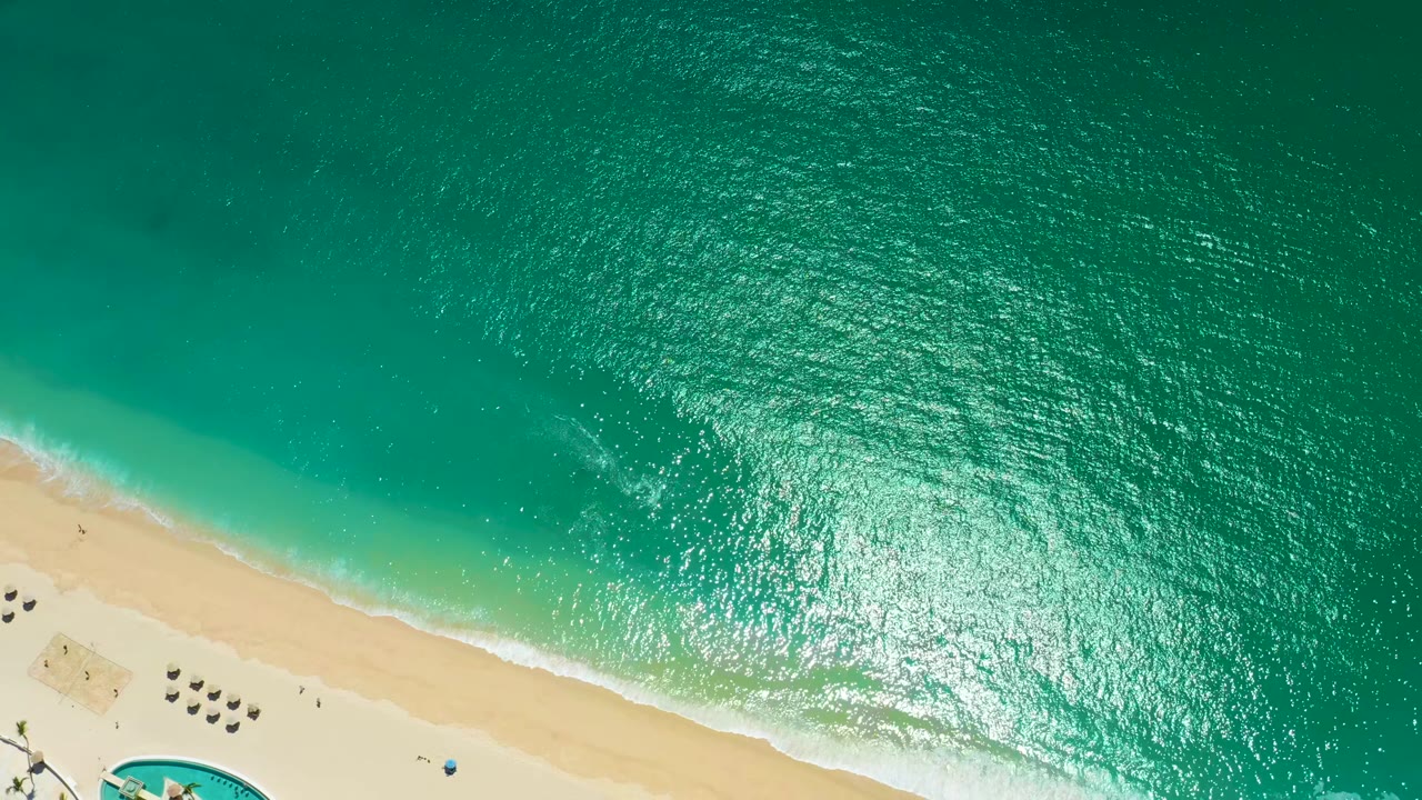 Overhead Marquis Los Cabos beach view with a larger patch of sun glare on the turquoise water, a thin white wave edge on the sand, scattered umbrellas near the beach, and no visible people.