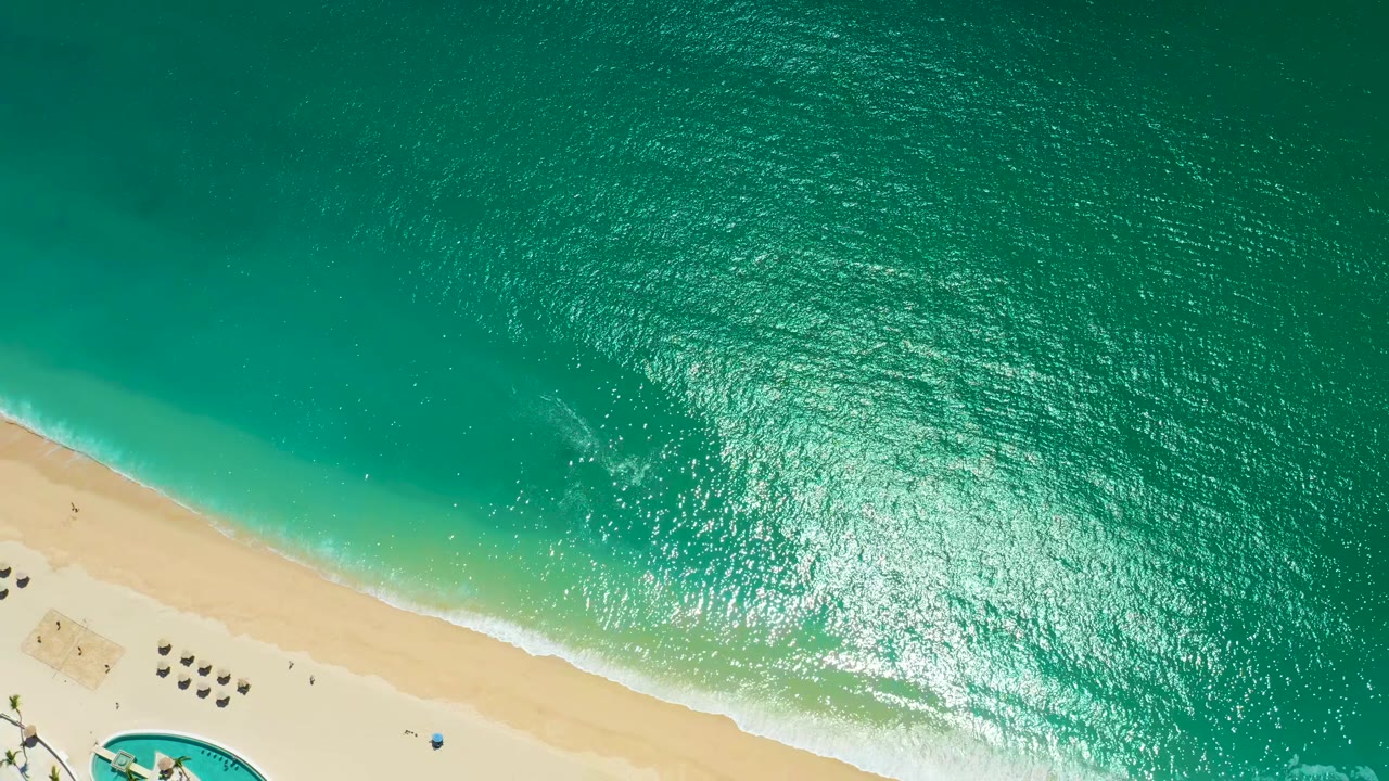 Overhead frame at Marquis Los Cabos, Mexico, with textured turquoise water dominating the image, the sandy beach crossing the lower left corner, broken white foam near shore, and no visible people.