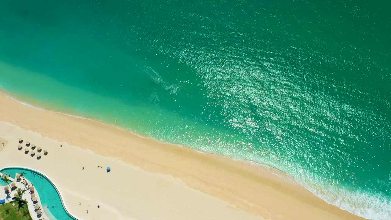 Opening aerial frame at Marquis Los Cabos, Mexico, showing turquoise water, a diagonal sandy shoreline, small beach umbrellas, a curved pool corner, and tiny people on the sand.