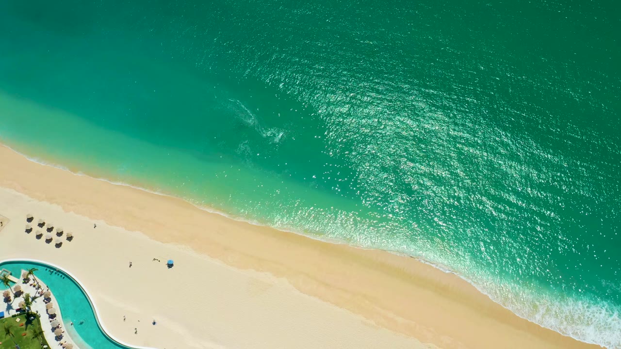 Top-down beach view with turquoise surf, pale sand, umbrellas, and a blue pool corner