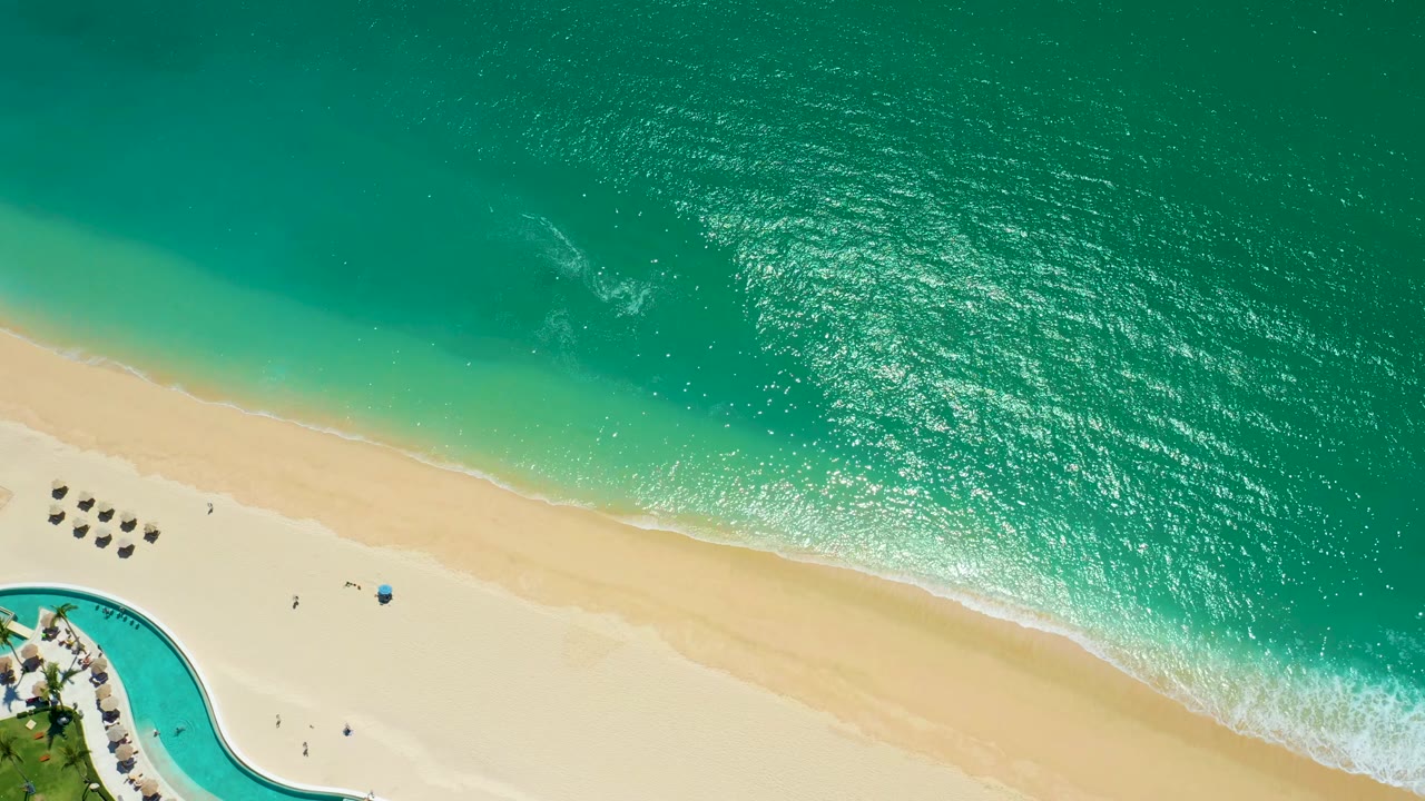 At 0:02, the aerial frame over Marquis Los Cabos shows a brighter patch of sunlit turquoise water above the beach, foam reaching the shore, rows of umbrellas, a pool curve, and tiny people below.