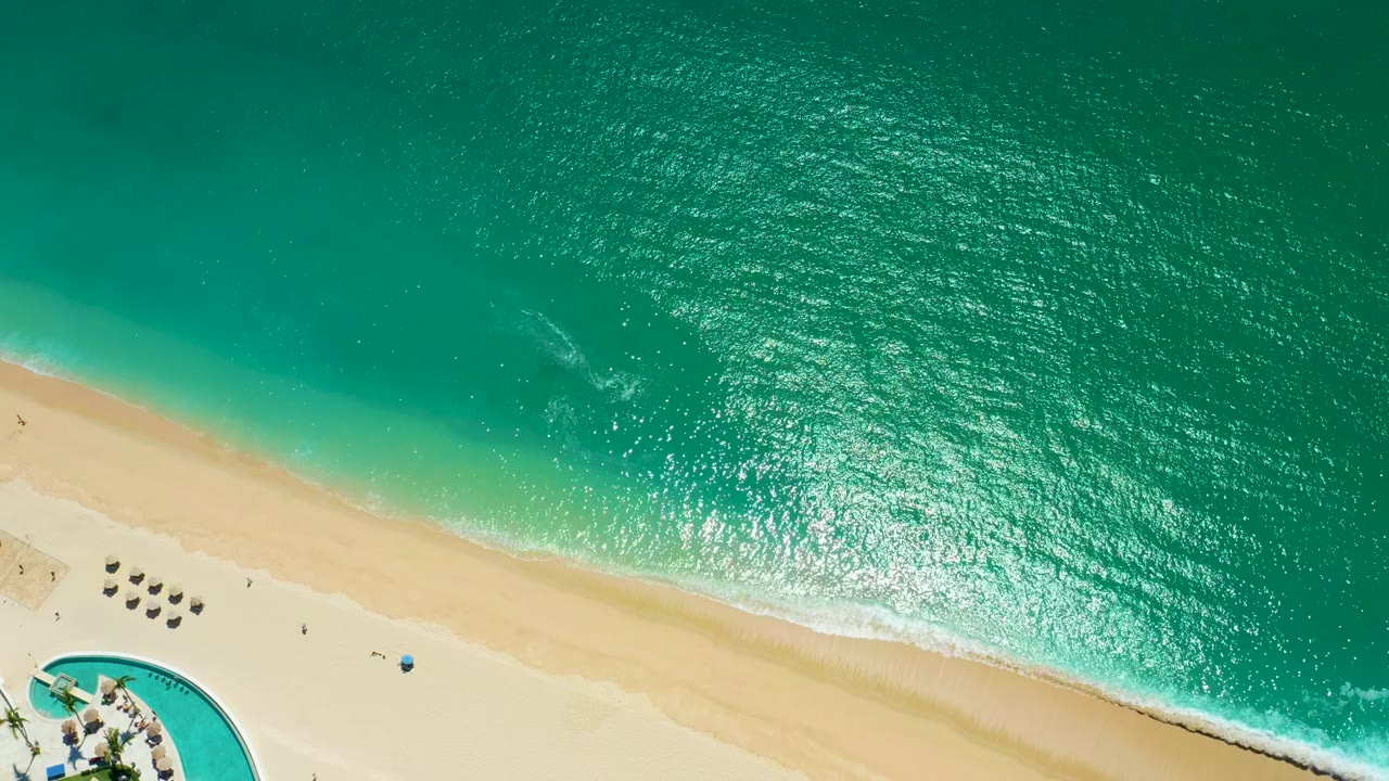 Overhead view at Marquis Los Cabos, Mexico, with turquoise ocean above a pale sandy beach, small surf lines, a curved pool edge, umbrellas, lounge chairs, and tiny people visible.