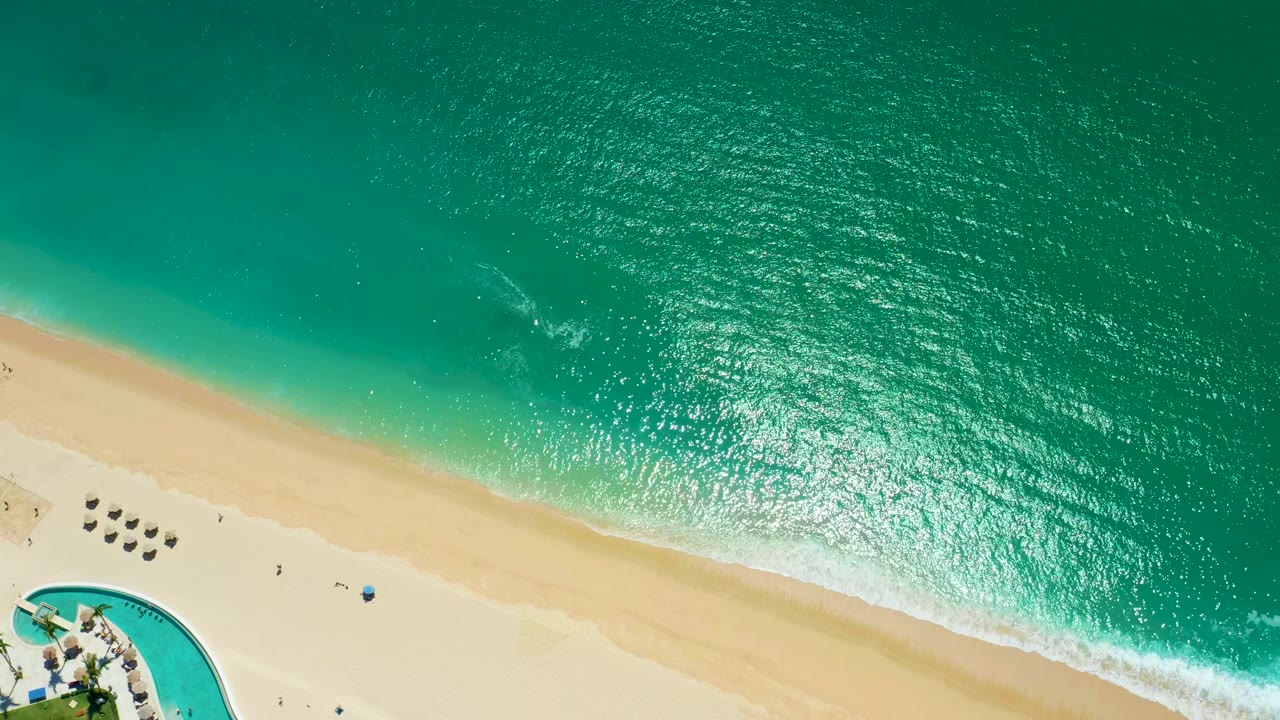 Top-down view at Marquis Los Cabos, Mexico, with sunlit turquoise ocean, pale sand, a thin white wave edge, grouped umbrellas, lounge chairs, pool curve, and tiny people visible.