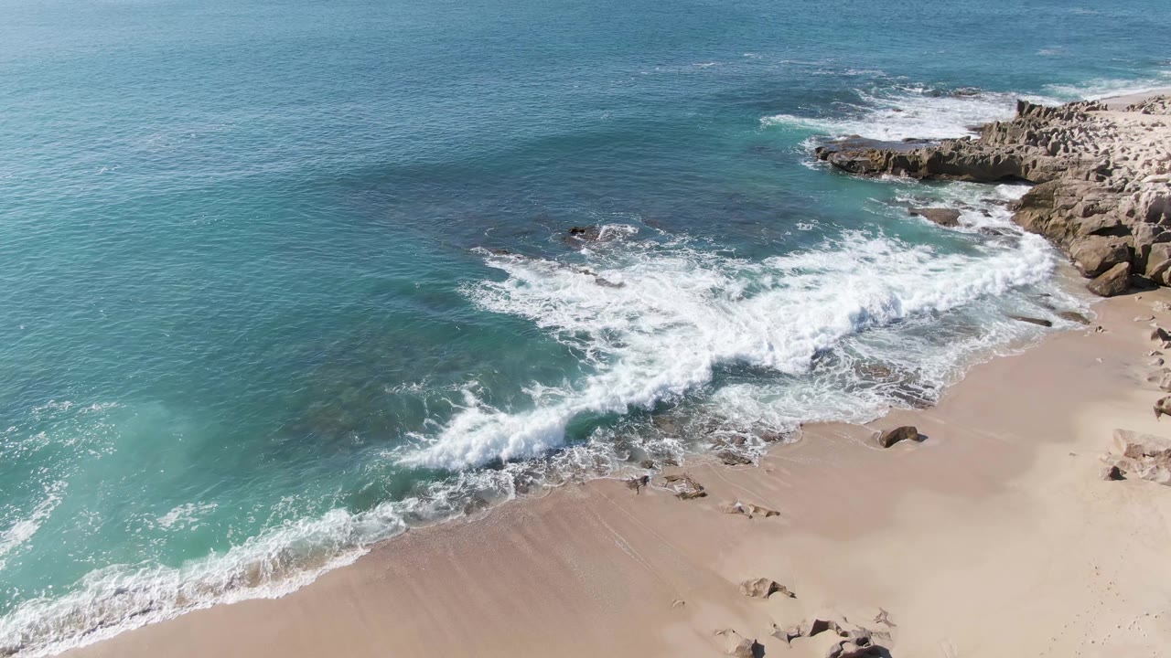Aerial view of waves breaking over sand and rocks beside turquoise ocean.