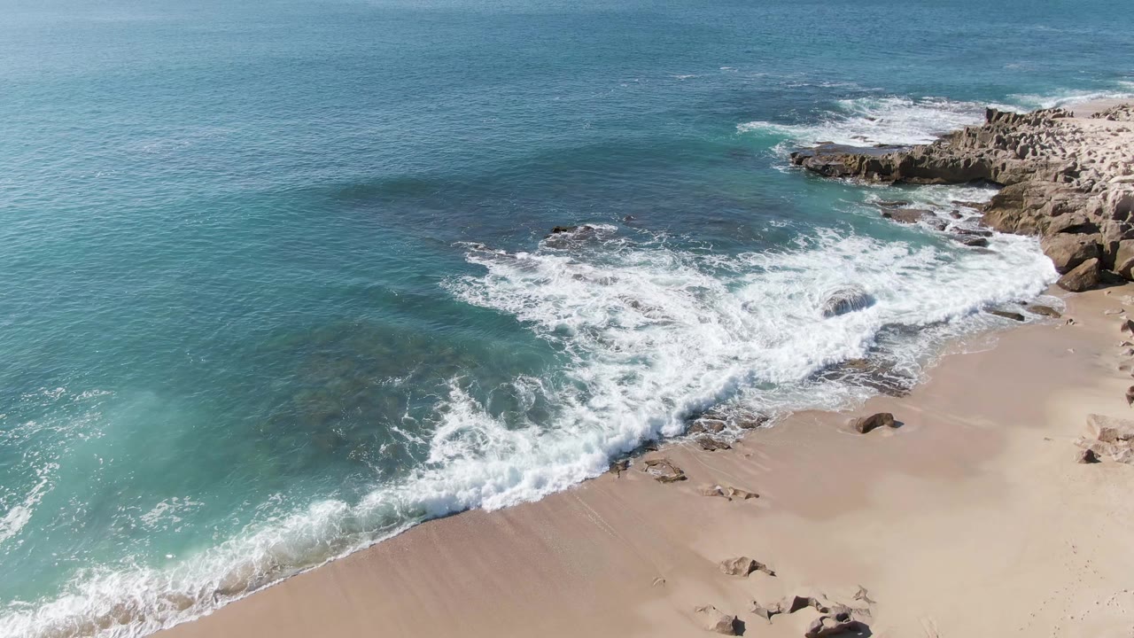 At 0:01, whitewater advances across the pale sand at Punta Gorda Beach while darker submerged rocks show through the turquoise shallows, with no visible people.