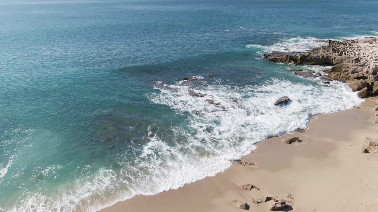 At 0:02, a broad band of foam curves along Punta Gorda Beach below the rocky right-hand point and open blue water, with no visible people.